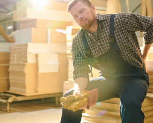 Tired manual worker with closed eyes sitting on stack of wooden plank and massaging lower back while feeling pain in back